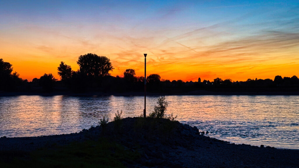 Rheinansicht bei Sonnenuntergang Rheinansicht bei Sonnenuntergang mit Schatten auf beiden Flussseiten. Der Himmel ist in verschiedenen Orangenen schattiert.