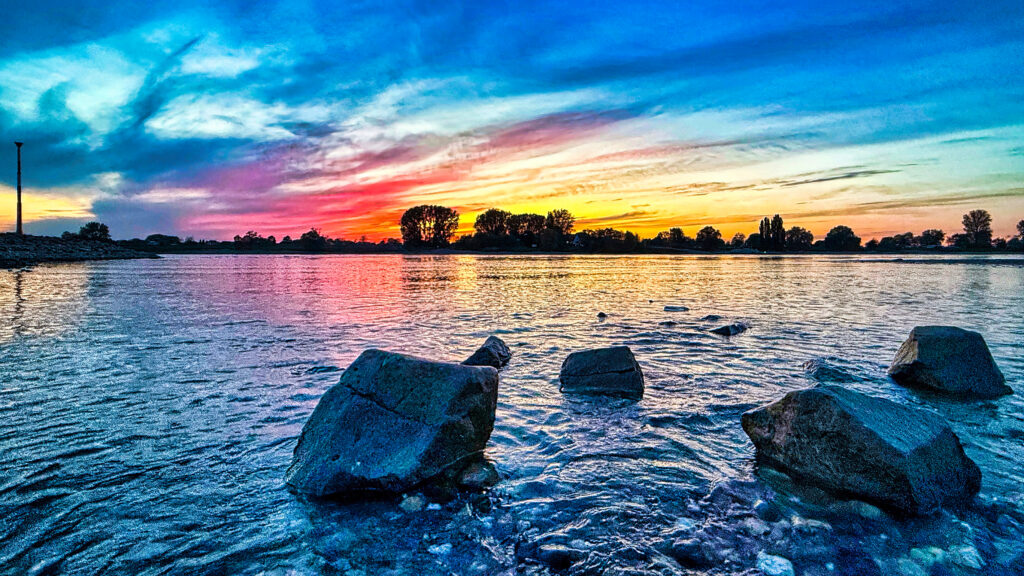 Rheinansicht bei Sonnenuntergang Rheinansicht in der Nähe des Fähranlegers in Blautönen und Himmel in Gelb- und Orangetönen. Im Vordergrund liegen große Steine im Wasser des Rheins.