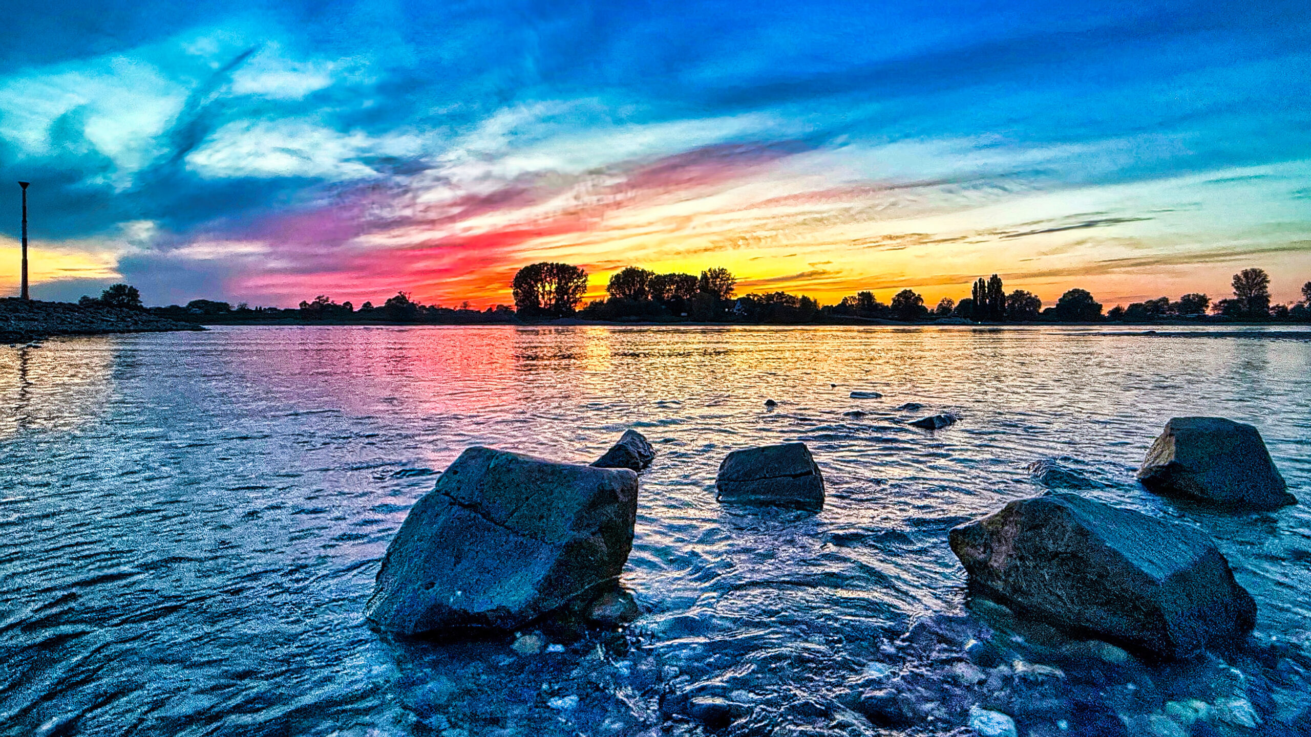Rheinansicht nĂ€he FĂ€hranleger Rheinansicht in der NĂ€he des FĂ€hranlegers in Blautönen und Himmel in Gelb- und Orangetönen. Im Vordergrund liegen groĂe Steine im Wasser des Rheins.
